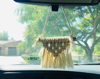 May include: A white macrame car hanging ornament with a wooden dowel and fringe. The ornament is hanging from the rearview mirror of a car.
