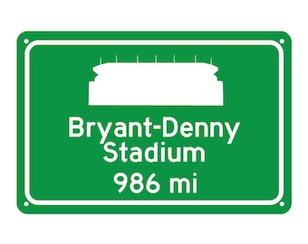 May include: A green road sign with white text and a white silhouette of a stadium. The sign reads "Bryant-Denny Stadium 986 mi".