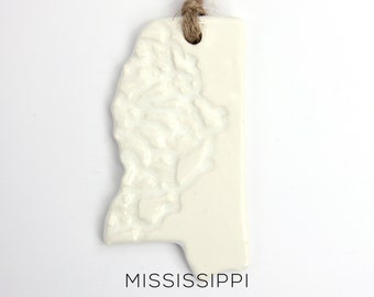 May include: White ceramic ornament shaped like the state of Mississippi with a textured surface and a twine hanger. The ornament has the word "MISSISSIPPI" printed below the state outline.