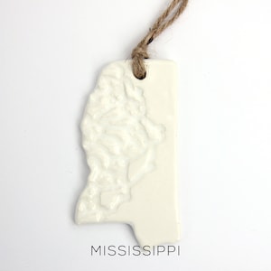 May include: White ceramic ornament shaped like the state of Mississippi with a textured surface and a twine hanger. The ornament has the word "MISSISSIPPI" printed below the state outline.