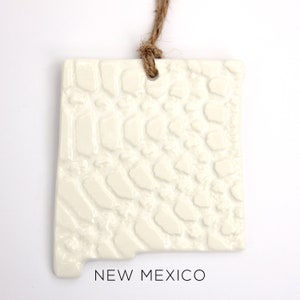 Puede incluir: Adorno de cerámica blanca con forma del estado de Nuevo México con una superficie texturizada. El adorno tiene un agujero en la parte superior para colgar y dice "NEW MEXICO" en la parte inferior.