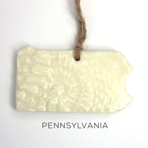May include: A white ceramic ornament shaped like the state of Pennsylvania with a twine hanger. The ornament has a textured surface and the word "PENNSYLVANIA" is printed below it.