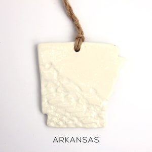 May include: White ceramic ornament shaped like the state of Arkansas with a twine hanger. The ornament has a textured surface and the word "ARKANSAS" is printed below the state.