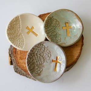 May include: Three small, round ceramic dishes with gold crosses. One is cream-coloured, another is a pale green, and the third is grey. Each dish has a textured lace pattern. The dishes are arranged on a wooden slice.