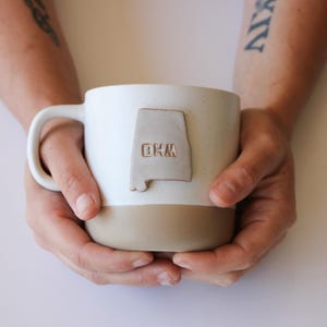 Peut inclure: Une tasse en céramique blanche avec une anse marron. La tasse a une bande marron autour du bas et un contour de l'Alabama en brun avec les lettres "BHM" dessus.