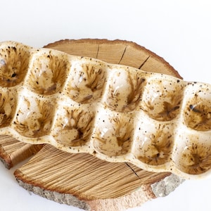 May include: A ceramic egg holder with twelve individual compartments. The holder is off-white with brown speckled detailing. It sits on a round, wooden slice with visible grain and bark.