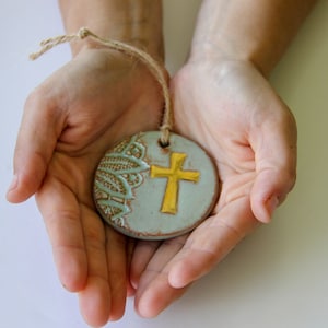 Pode incluir: Um ornamento redondo de cerâmica com fundo azul claro e cruz dourada. O ornamento tem um padrão de renda decorativo no lado esquerdo e um fio de juta para pendurar. O ornamento é segurado em mãos abertas.