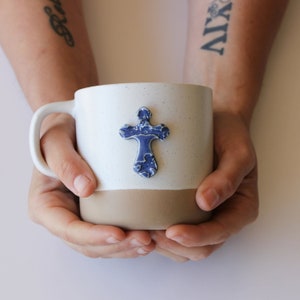 May include: A white ceramic mug with a brown rim and a blue ceramic cross attached to the front. The mug is being held by two hands.