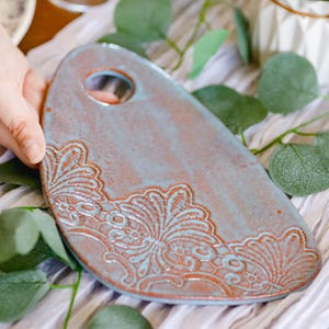 Pode incluir: Tábua de servir de cerâmica feita à mão com formato único e furo para pendurar. A tábua apresenta um esmalte azul-esverdeado com um detalhado desenho floral marrom-avermelhado. A tábua tem aproximadamente 30 cm de comprimento.