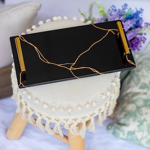 May include: A rectangular black tray with gold handles and gold vein-like accents. The tray is resting on a small, round, white stool with a cream-colored, fringed skirt. Decorative home decor item.