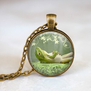 May include: A round pendant necklace with a bronze-colored chain. The pendant features a green frog relaxing on a bed of grass against a green background with floral accents. The frog is the focal point of the pendant.