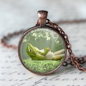 May include: A round pendant necklace with a copper-colored chain. The pendant features a green frog relaxing on a lily pad, set against a green background with floral accents. The frog is the focal point of the pendant.