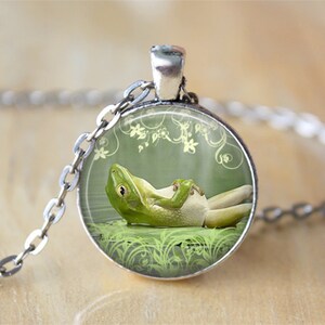 May include: A silver-toned necklace featuring a round pendant with a green frog illustration. The frog is depicted relaxing in water, with a light green background and floral accents. The chain is a classic link style.