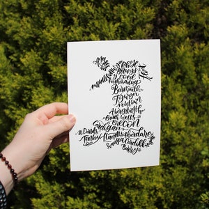 May include: A white card with black calligraphy listing Welsh towns and cities. The towns and cities are arranged in a shape resembling a map of Wales.