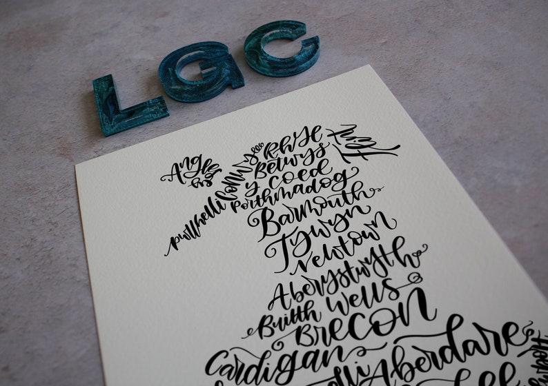 May include: A black and white print of the Welsh flag, with the names of Welsh towns and cities written in a calligraphic style. The names include: Anglesey, Brecon, Caernarfon, Cardigan, Carmarthen, Conwy, Denbigh, Gwynedd, Llandudno, Llanelli, Merthyr Tydfil, Monmouthshire, Newport, Pembrokeshire, Powys, Rhondda Cynon Taff, Swansea, Vale of Glamorgan, Wrexham.