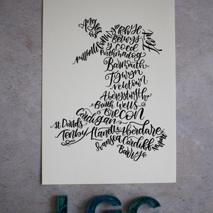 May include: A white card with black calligraphy listing towns and cities in Wales. The text is arranged in the shape of the country of Wales. The card is sitting on a gray surface with three teal blue letters that spell out "LGC".