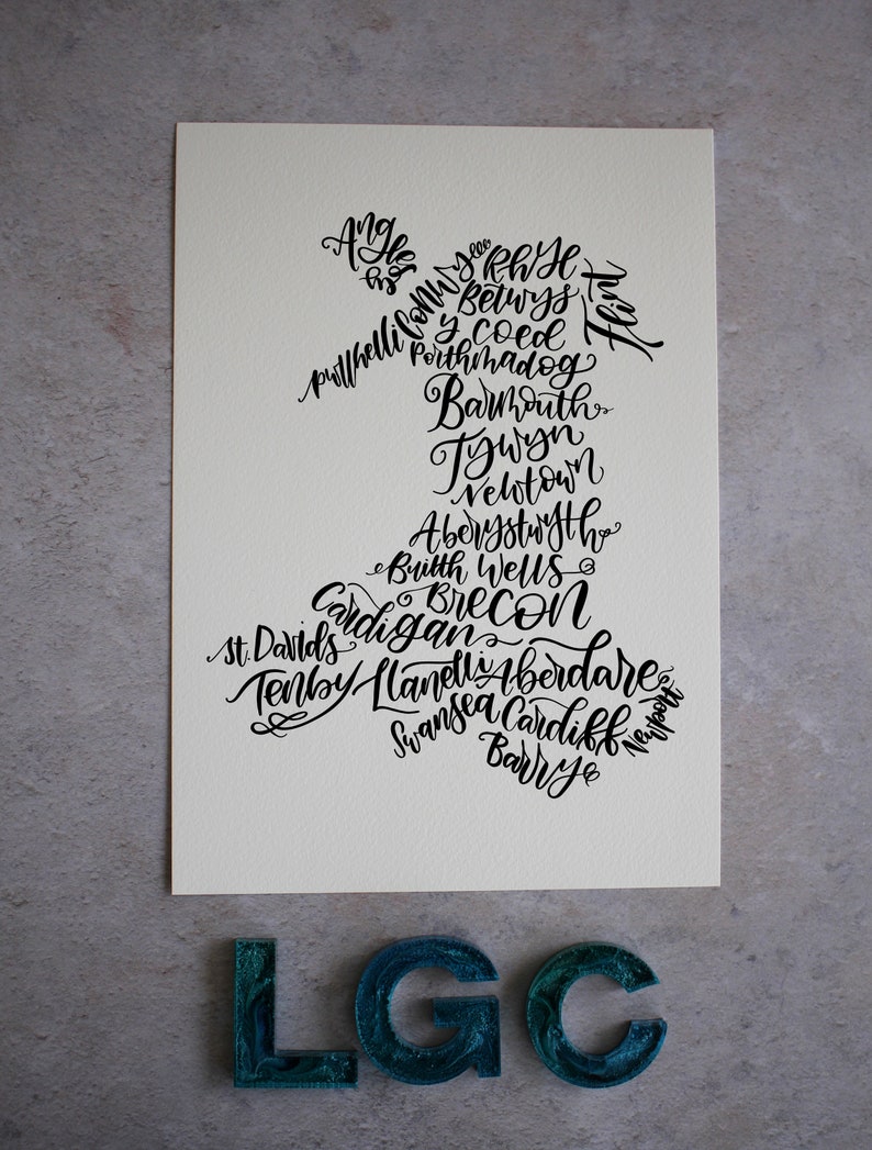 May include: A white card with black calligraphy listing towns and cities in Wales. The text is arranged in the shape of the country of Wales. The card is sitting on a gray surface with three teal blue letters that spell out "LGC".