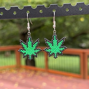 May include: A pair of dangle earrings featuring a beaded leaf design. Each earring is crafted from small, square-shaped green beads outlined with black beads, suspended from silver-toned metal fishhook wires for pierced ears.