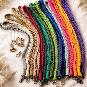 May include: A collection of braided lanyards in various colors, including gray, tan, red, green, blue, and purple. Each lanyard has a metal clasp and loop. The lanyards are arranged on a white surface with dried plants in the background.