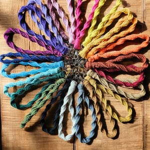 May include: A collection of colorful macrame lanyards in various shades, including purple, pink, yellow, orange, and blue. Each lanyard is twisted and attached to a metal clasp. The lanyards are arranged in a circular pattern on a wooden surface.