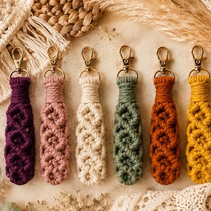 May include: Six macrame keychains arranged in a row, each featuring a gold-toned lobster clasp and a knotted cord design. The colors from left to right are deep purple, dusty rose, cream, sage green, burnt orange, and mustard yellow.