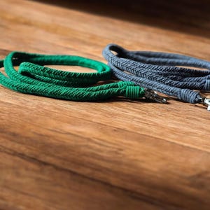 Op de afbeelding: Twee gevlochten touwkoorden in groen en grijs, elk met een metalen sluiting. De koorden liggen op een houten oppervlak. Het groene koord heeft een levendige, effen kleur, terwijl het grijze een getextureerde uitstraling heeft.