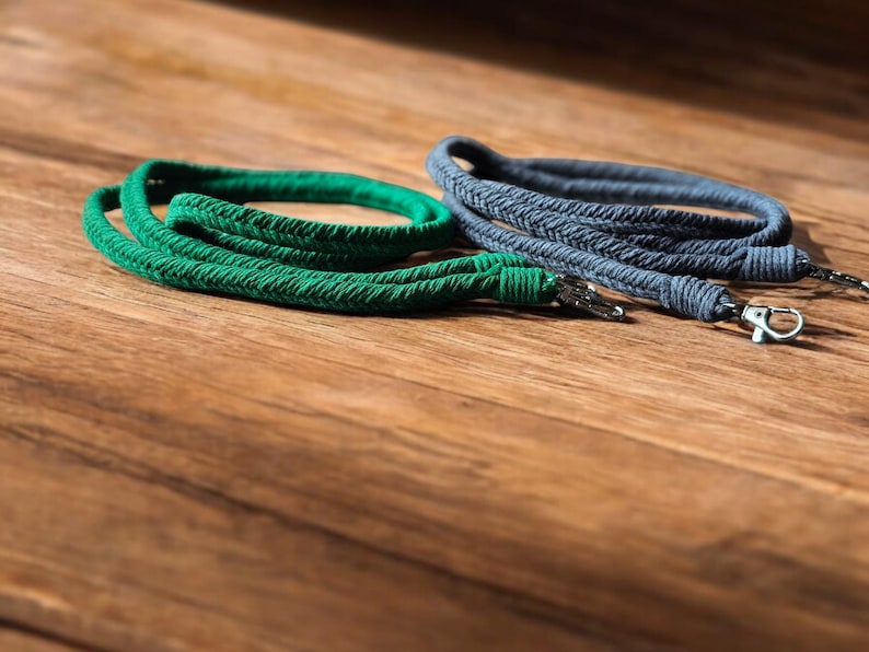 Op de afbeelding: Twee gevlochten touwkoorden in groen en grijs, elk met een metalen sluiting. De koorden liggen op een houten oppervlak. Het groene koord heeft een levendige, effen kleur, terwijl het grijze een getextureerde uitstraling heeft.