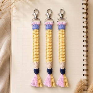 May include: Three macrame keychains shaped like pencils, each featuring a silver lobster clasp, a pink top section, a light blue band, a long yellow body, a beige tip, a dark blue knot, and a tassel with pink and yellow cotton cord strands.