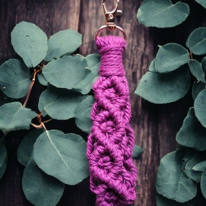 May include: A handmade macrame keychain in a vibrant magenta color. The keychain features a woven design and a gold-colored clasp. The background includes green leaves and a dark wooden surface.