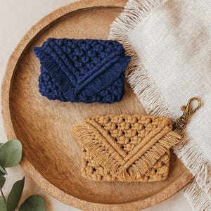 May include: Two macrame wallets, one navy blue and one tan, rest on a wooden tray. The wallets have a woven design with fringe detailing. A brass keychain is attached to the tan wallet. A white fringed cloth is next to the wallets.