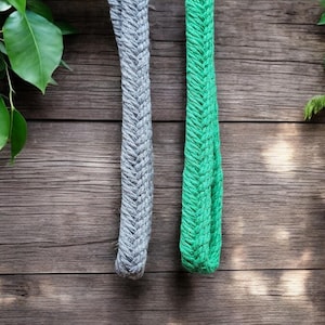 Op de afbeelding: Twee gevlochten touwen, &eacute;&eacute;n grijs en &eacute;&eacute;n groen, hangen tegen een rustieke houten achtergrond. De touwen hebben een textuur, geweven uiterlijk. Groene bladeren omlijsten de boven- en rechterkant van de afbeelding en voegen een natuurlijk element toe.