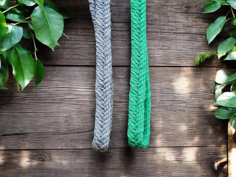 Op de afbeelding: Twee gevlochten touwen, &eacute;&eacute;n grijs en &eacute;&eacute;n groen, hangen tegen een rustieke houten achtergrond. De touwen hebben een textuur, geweven uiterlijk. Groene bladeren omlijsten de boven- en rechterkant van de afbeelding en voegen een natuurlijk element toe.