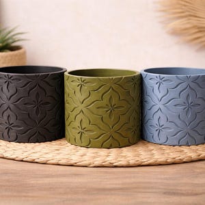 May include: Three cylindrical plant pots in black, olive green, and slate blue. Each pot features a repeating embossed floral pattern across its exterior surface. The pots are arranged in a horizontal row, showcasing a consistent design and shape.