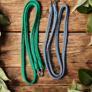 Op de afbeelding: Twee gevlochten lanyards, &eacute;&eacute;n smaragdgroen en &eacute;&eacute;n lichtblauw, met zilverkleurige metalen draaihaken. De lanyards worden gepresenteerd op een rustiek houten oppervlak, met groene bladeren op de achtergrond.