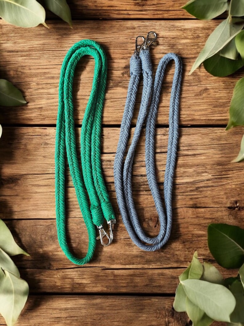 Op de afbeelding: Twee gevlochten lanyards, &eacute;&eacute;n smaragdgroen en &eacute;&eacute;n lichtblauw, met zilverkleurige metalen draaihaken. De lanyards worden gepresenteerd op een rustiek houten oppervlak, met groene bladeren op de achtergrond.