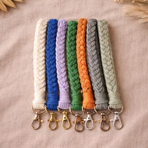 May include: A collection of seven braided lanyards in various colors, including cream, blue, lavender, green, orange, gray, and sage green. Each lanyard has a gold or silver-colored clasp. The lanyards are arranged on a neutral-toned fabric.