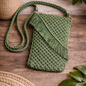 May include: A green crocheted crossbody bag with a long braided strap. The bag features a flap with fringe and gold-colored hardware. The bag is made of a textured, woven material and is displayed on a wooden surface.