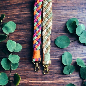 May include: Two braided rope straps with brass-colored clasps. One strap features a vibrant mix of pink, orange, purple, and green. The other strap is a blend of beige, white, and green. The straps are displayed on a dark wood surface with eucalyptus leaves.