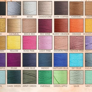 Op de afbeelding: Een kleurenkaart met een verscheidenheid aan garenkleuren. De kaart toont vierkanten garen in tinten als zwart, grijs, beige en zandsteen. Andere kleuren zijn kaki, mosterd en verschillende tinten roze, paars en groen.