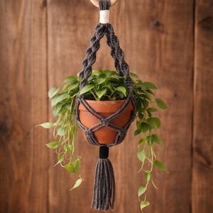 May include: A hanging macrame plant holder with a terracotta pot filled with green plants. The macrame is dark gray, with a wooden ring at the top and a tassel at the bottom. The background is a wooden wall.