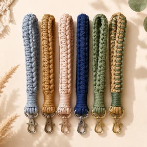 May include: Six macrame wristlet lanyards in various colors including light blue, mustard yellow, pale pink, navy blue, sage green, and a two-tone green and tan. Each lanyard features a braided cord design and a metal lobster claw clasp.