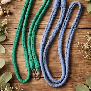 May include: Two rope lanyards, one green and one blue, with silver-colored metal clasps. The lanyards are made of a thick, braided rope material. The background is a wooden surface with decorative greenery.