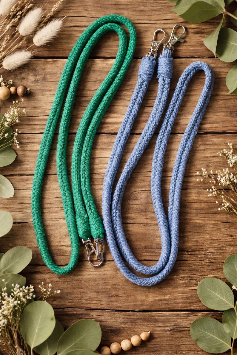 Op de afbeelding: Twee touw lanyards, &eacute;&eacute;n groen en &eacute;&eacute;n blauw, met zilverkleurige metalen sluitingen. De lanyards zijn gemaakt van een dik, gevlochten touwmateriaal. De achtergrond is een houten oppervlak met decoratief groen.