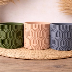 May include: Three cylindrical planters in olive green, peach, and slate blue, each with a repeating arched pattern. They sit on a round woven mat, with decorative vases and dried plants in the background.