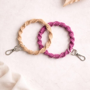 May include: Two macrame wrist keychains. One is tan, the other is purple. Each keychain has a silver-colored clasp. The keychains are circular and have a twisted rope design. The background is white with a dried palm leaf.
