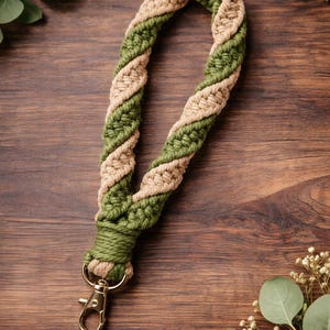 May include: A macrame wristlet in a twisted design, featuring alternating sections of olive green and tan yarn. The wristlet has a gold-colored clasp for attaching keys or other small items. The background is a dark wood surface.