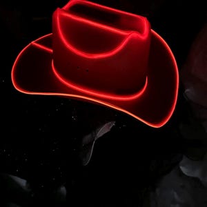 May include: A cowboy hat outlined with bright red glowing neon light piping. The hat features a structured crown and a curved brim, with the vibrant red illumination highlighting its silhouette against a dark, black background.