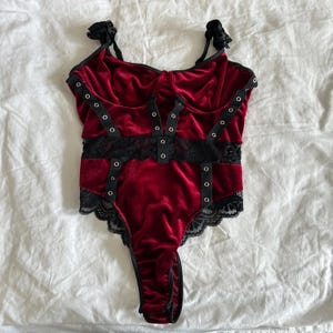 May include: A deep red velvet bodysuit featuring black lace trim along the waist and leg openings. The garment includes black straps with silver-toned metal grommets arranged in a vertical pattern across the front and along the shoulder straps.