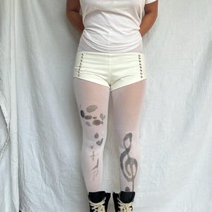 May include: Cream-colored shorts with silver eyelets and the word "UNREAL" printed on the side. The model is wearing white tights with gray abstract designs and a treble clef. Black high-top sneakers complete the look.