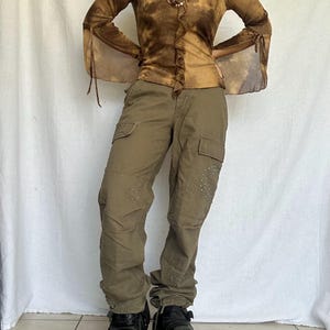 May include: A brown tie-dye long-sleeve top with bell sleeves and olive green cargo pants. The pants feature multiple pockets and studded heart designs. Black boots complete the outfit, creating a unique, stylish look.